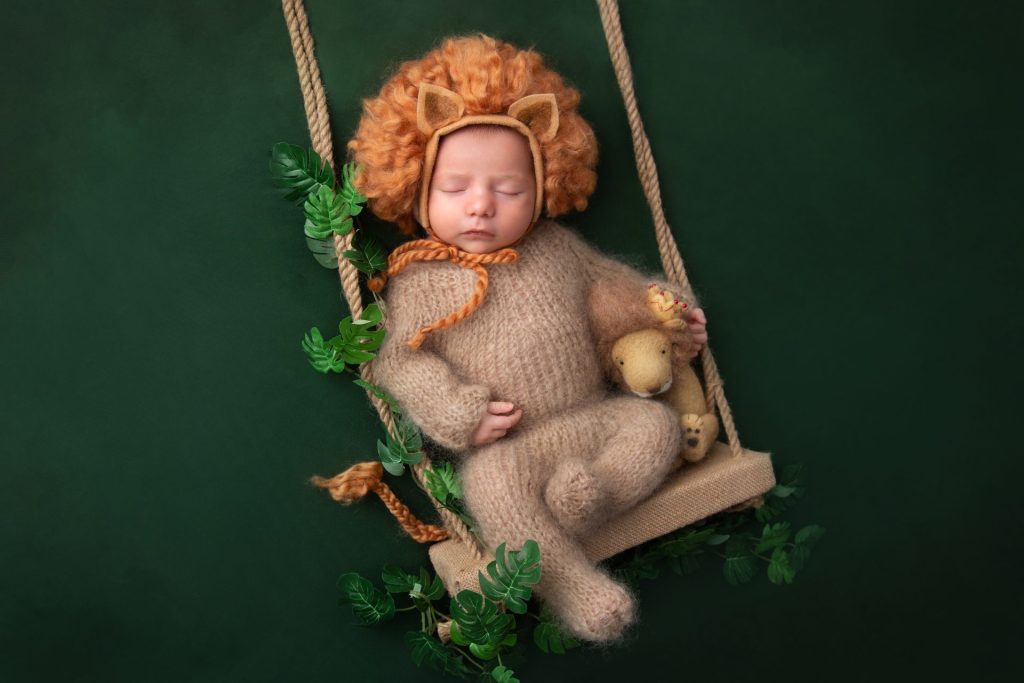 a baby boy dressed in a newborn lion outfit poses on a swing prop for newborn photos in nj
