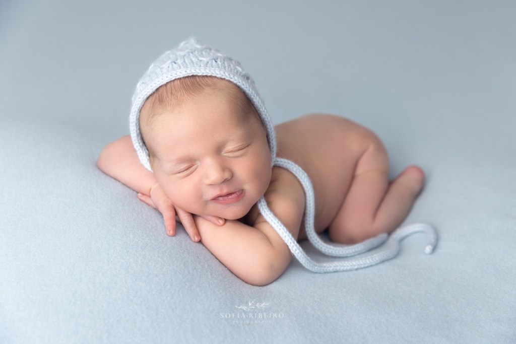 a chubby newborn boy smiles, posed in baby blue during his photo session in nj