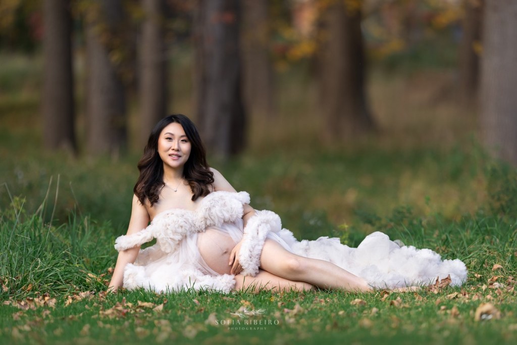 a mom poses for her maternity photo while lying in a field of grass in a white gown with fall trees in the background in nj