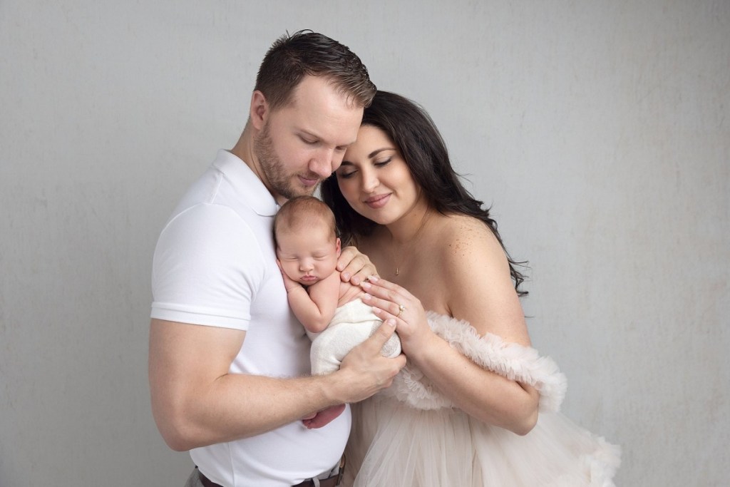 New parents sniggle with their sleeping newborn baby on dad's chest while standing in a studio after visiting baby classes nj