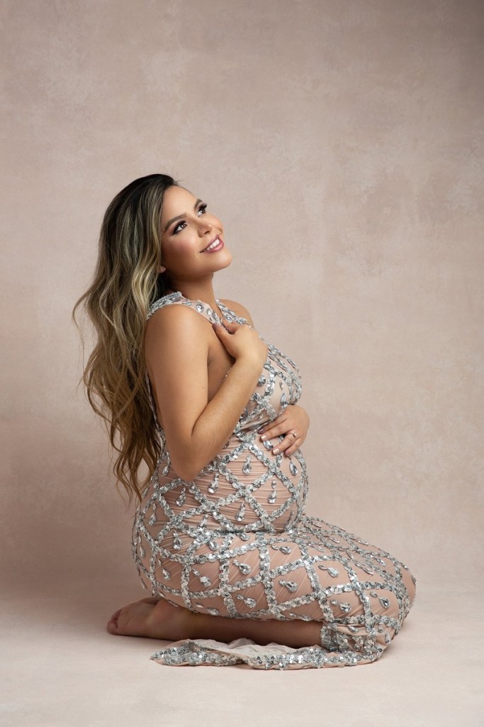 A mom to be in a maternity gown kneels on a studio floor with a hand on chest before visiting baby shower venues essex county