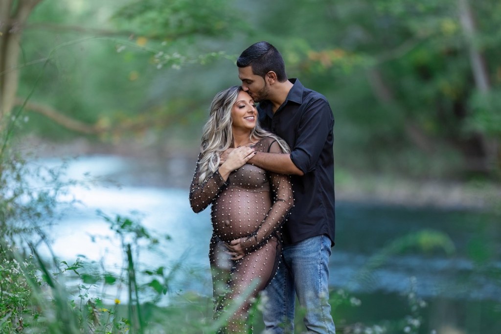 A mom to be in a sheer maternity outfit leans into and is kissed by her husband on the edge of a river