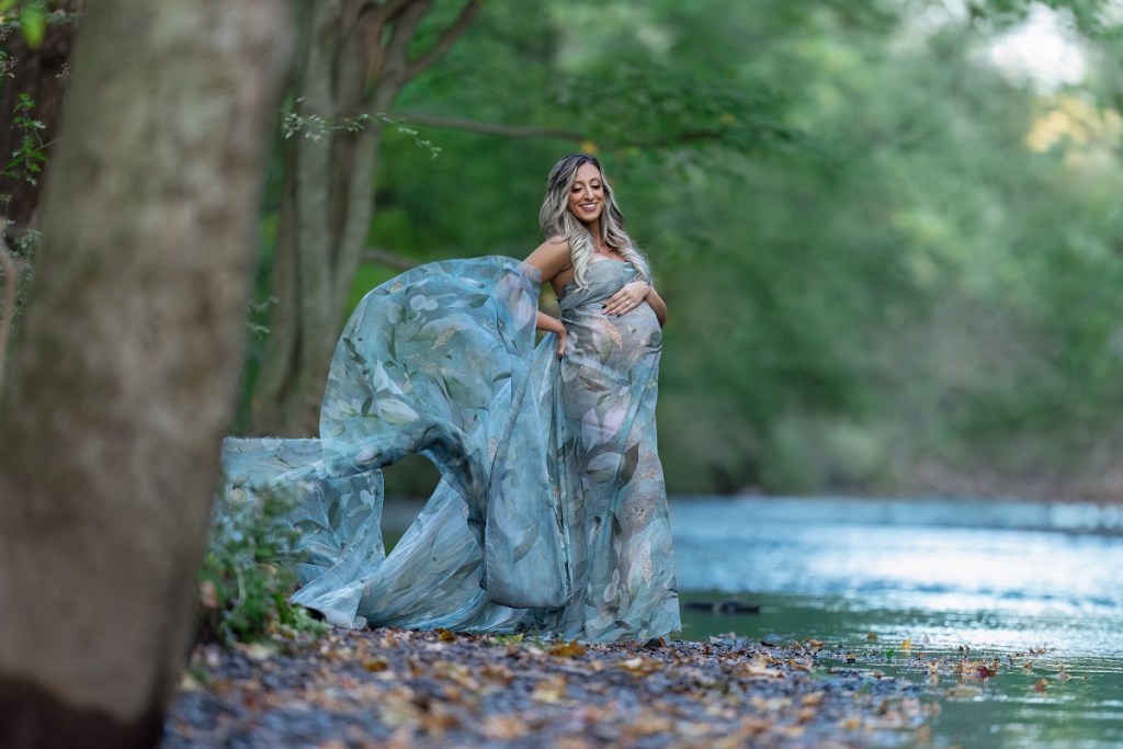A mom to be a blue flowing maternity gown stands on the edge of a river smiling down to her bump after meeting with midwives in new jersey