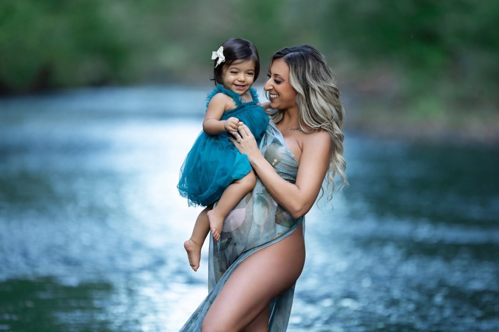 A pregnant mother in a blue maternity gown stands by a river with her toddler daughter on her bump after meeting with midwives in new jersey