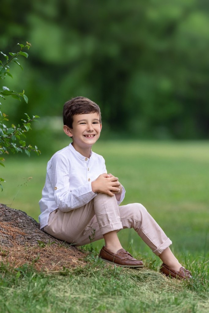 A young boy in a white shirt and khakis sits on a tree root in a park after meeting with nannies in new jersey