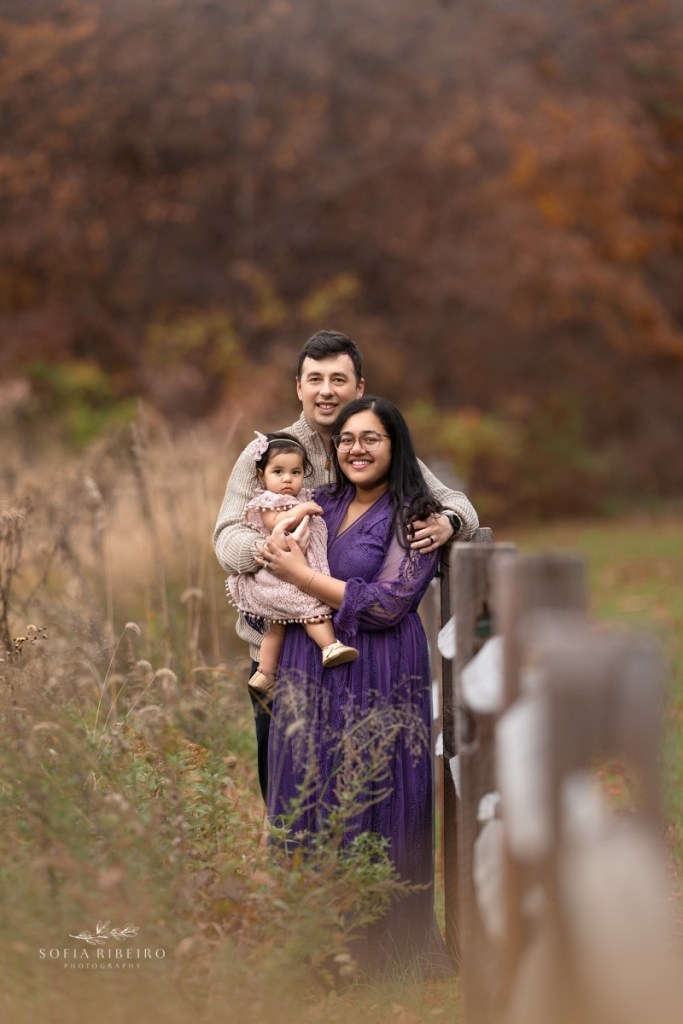 mom dad and baby girl along a wooden fence in a field before taking family photos in nj