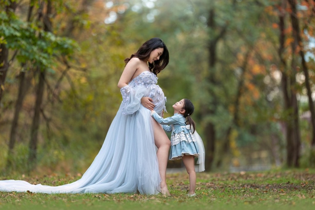 A pregnant mother stands in a park in a blue maternity gown as her toddler daughter leans into the bump after a prenatal massage new jersey
