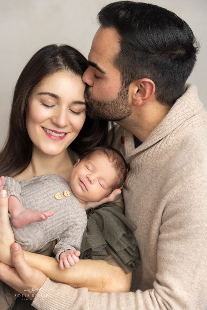 mom and dad cuddle newborn as he smiles in his sleep