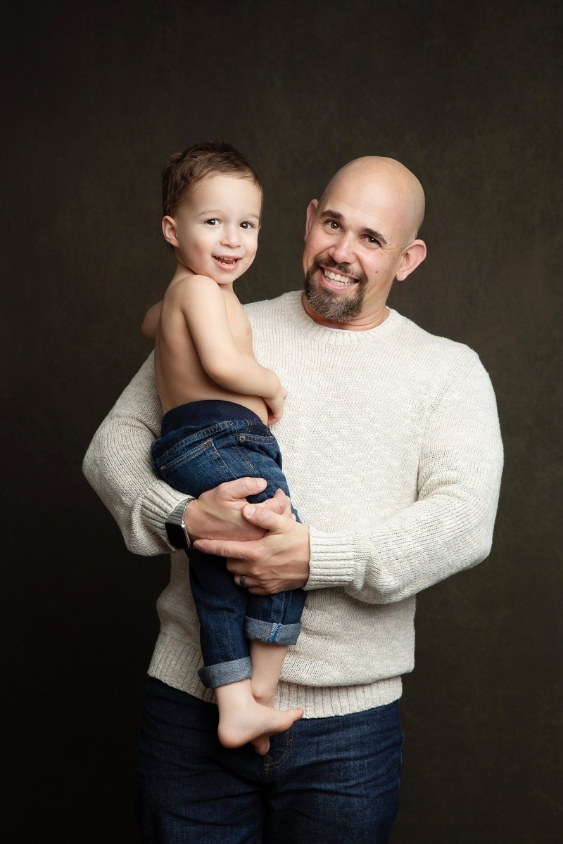 A happy father in a white sweater stands in a studio holding his toddler son with no shirt and jeans