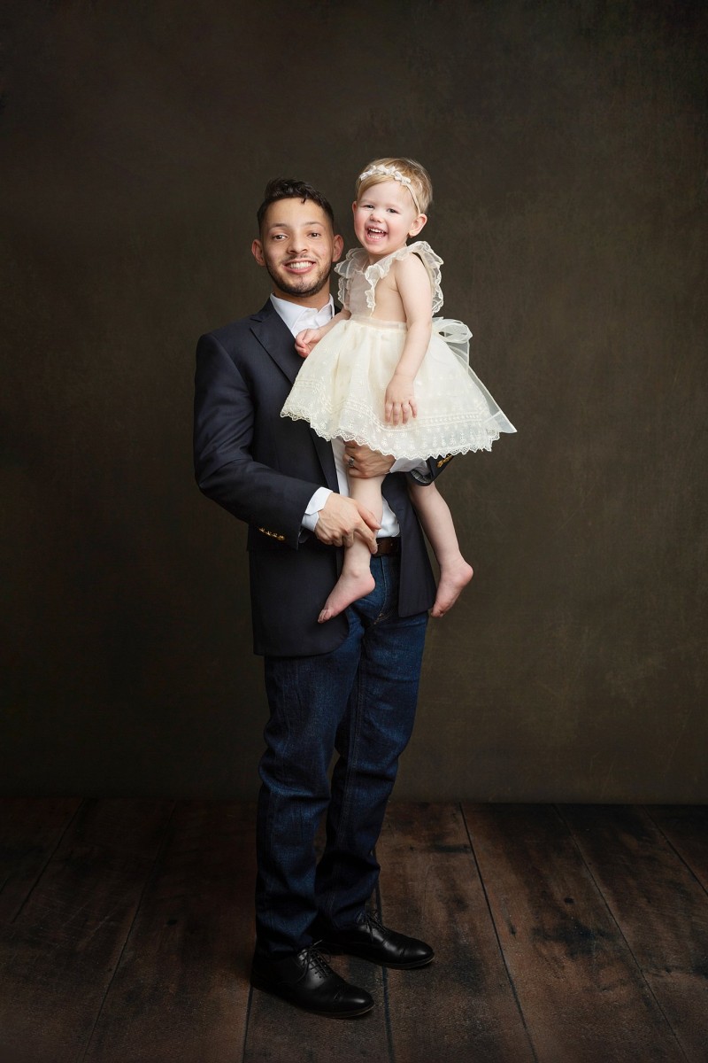 A happy father in a suit jacket stands in a studio holding his laughing toddler daughter after visiting dental family junior