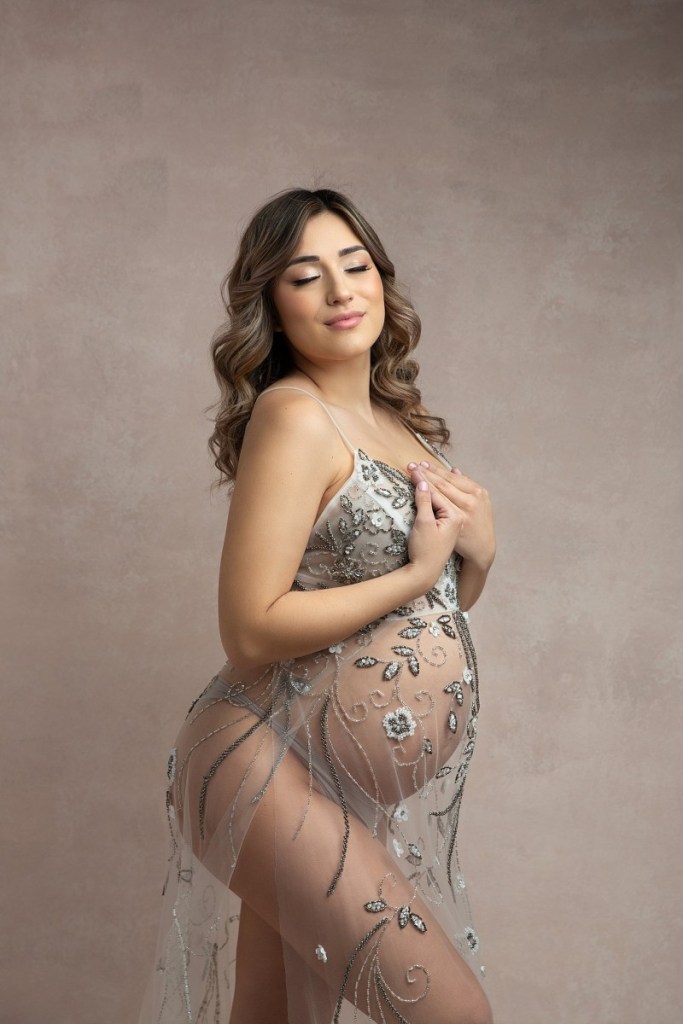 A mom to be stands in a studio with her hands on her chest in a sheer embroidered maternity gown