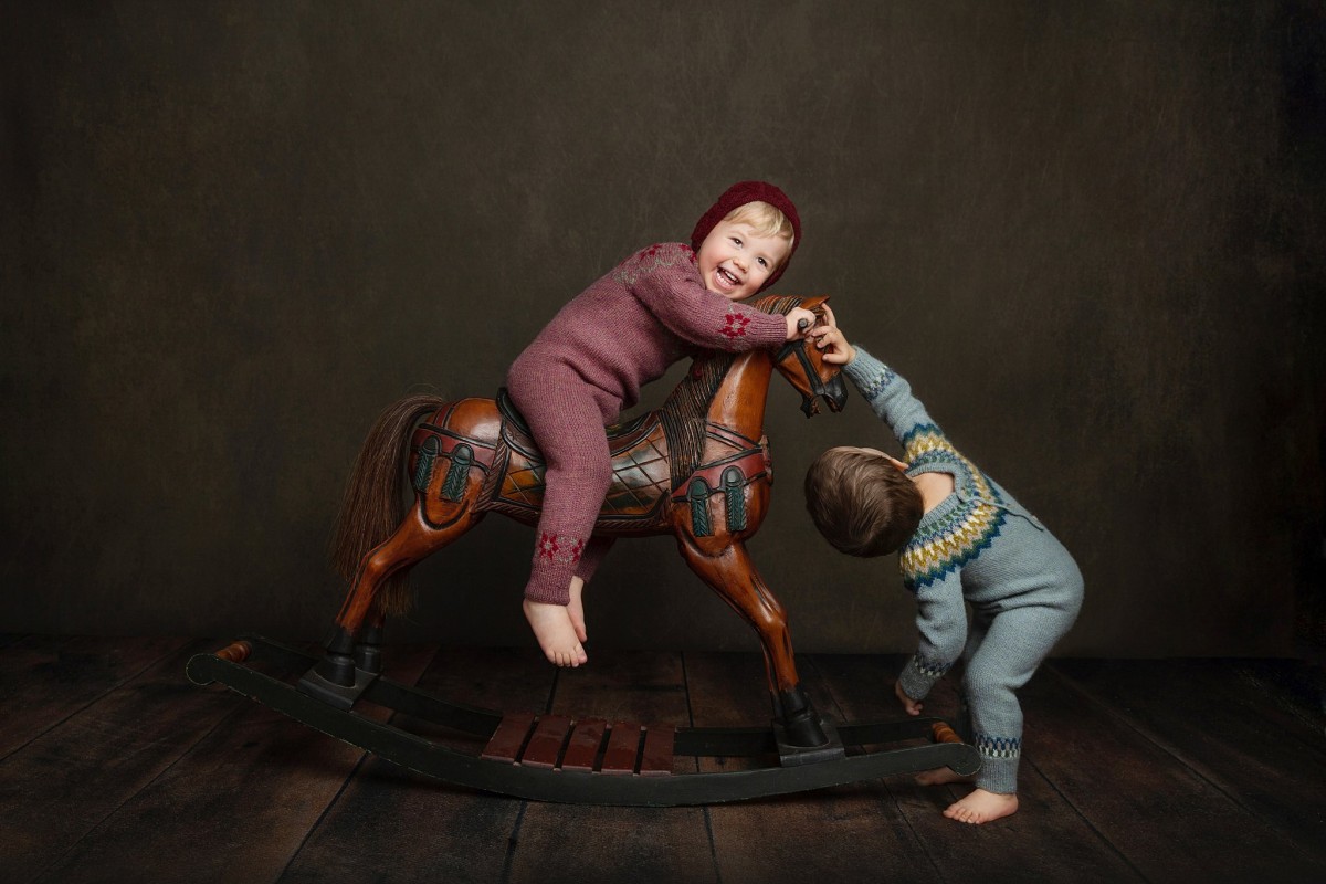 Toddler siblings in knit pajamas play on a wooden horse in a studio
