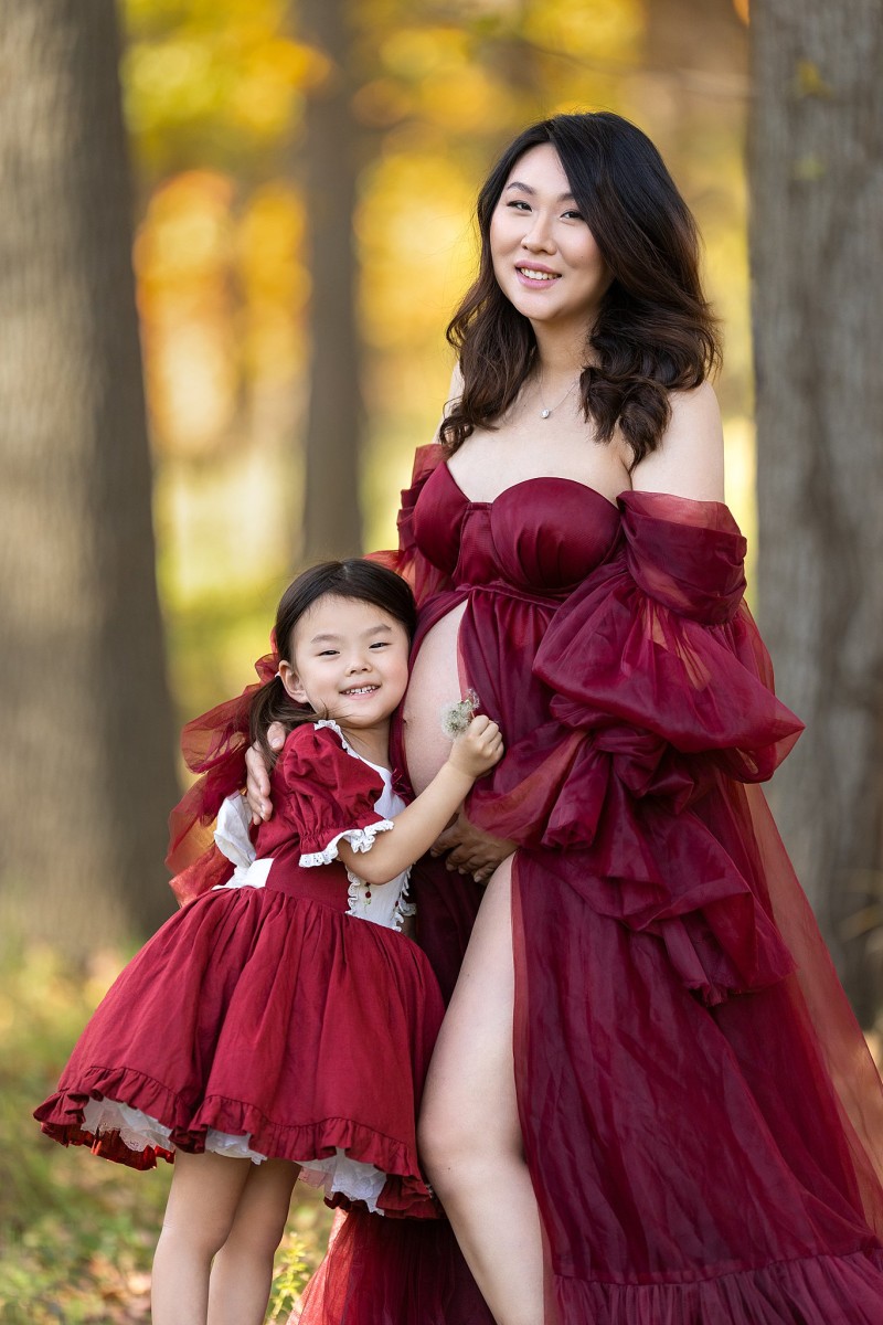 An expecting mother in a red tule maternity gown stands in a forest with her young daughter hugging the bump in a red dress