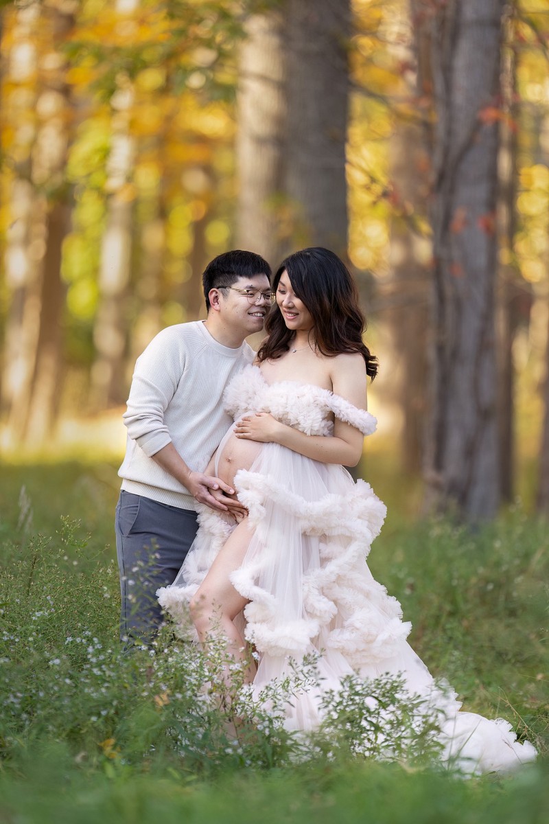 A mom to be stands in a forest in a white tule maternity gown while holding the bump and smiling with her husband after visiting new beginnings obgyn