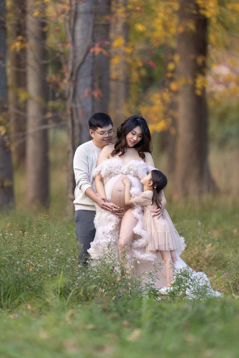 A mother to be stands in a forest in a white maternity gown with bump exposed with her husband and young daughter touching the bump after visiting new beginnings obgyn