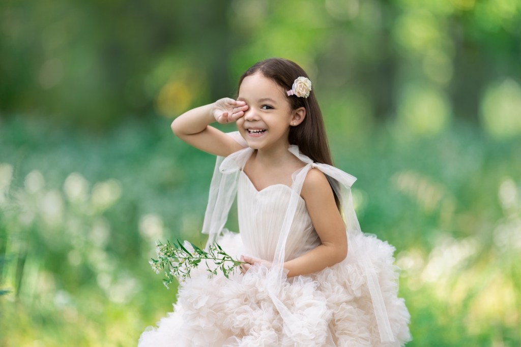 little girl playfully hides one eye while frolicking in a field of white flowers in a ballgown in nj during a portrait session with an nj photographer