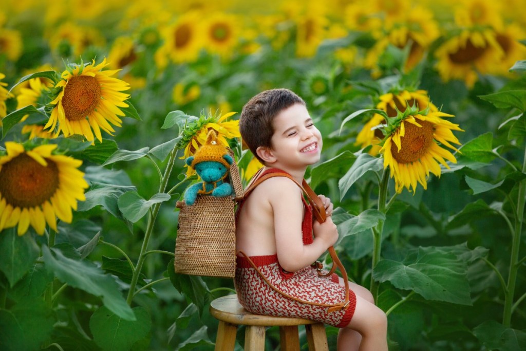 little boy smiles with a bear in his backpack field of sunflowers in handknit outfits while taking photos with a childrens photographer in new jersey