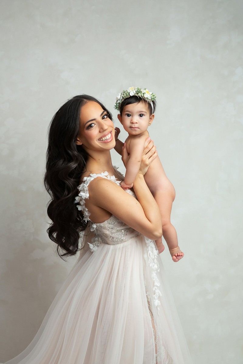 A mother in a white embroidered dress lifts her infant child while standing in a studio thanks to an obgyn westfield nj