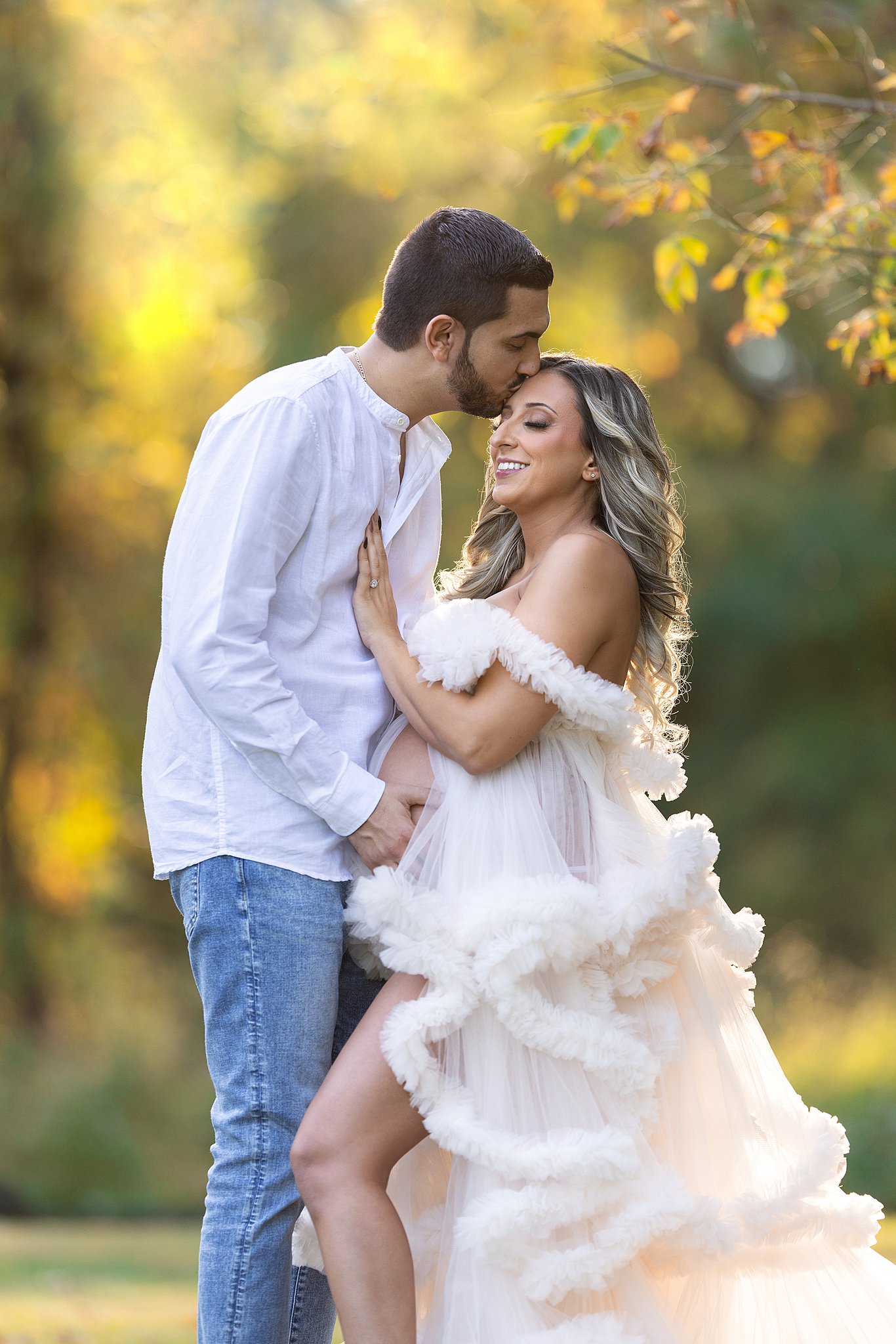 A father kisses his pregnant wife on the forehead while standing in a forest at sunset and holding the bump