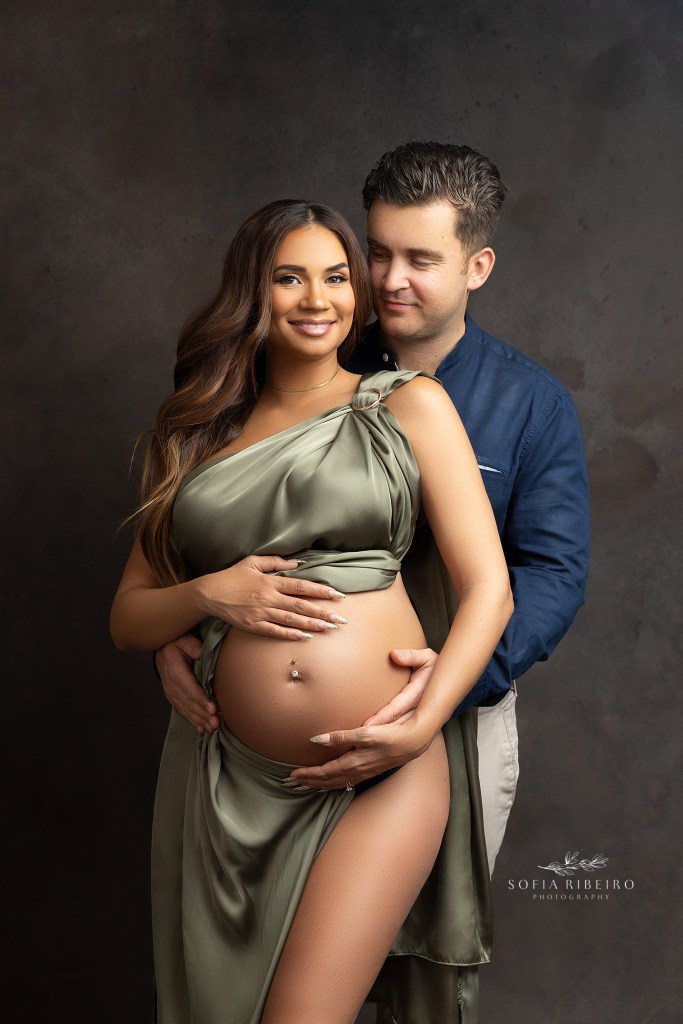 dad looks lovingly at mom as she smiles to the camera draped in green silk fabric, with a hoboken nj maternity photographer