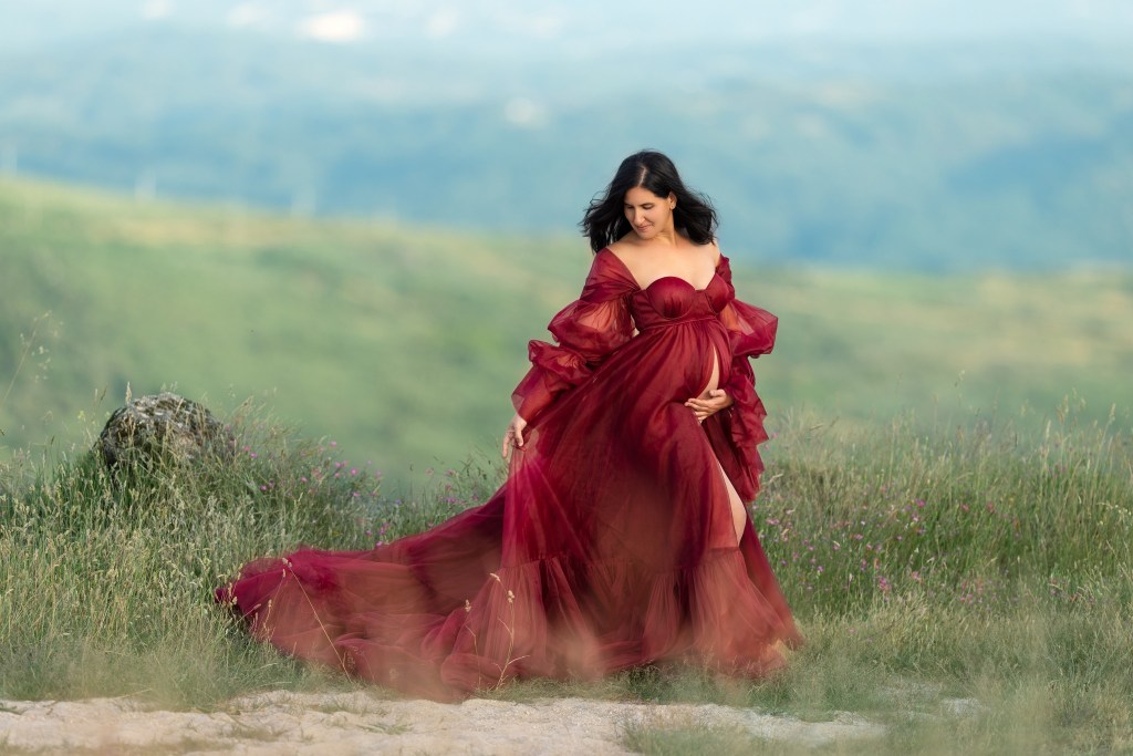 a mom poses in a red maternity gown outdoors with a mountain range in the background.