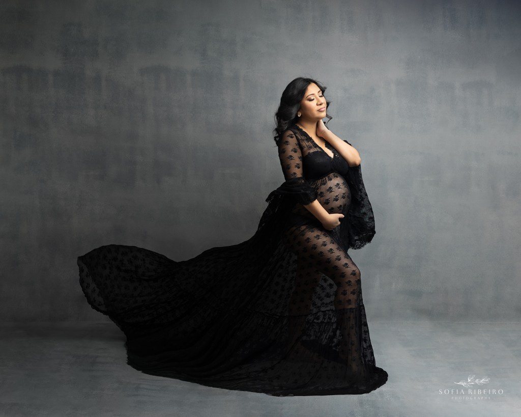 mom poses looking into the light, wearing a black lace dress during maternity portraits in new jersey
