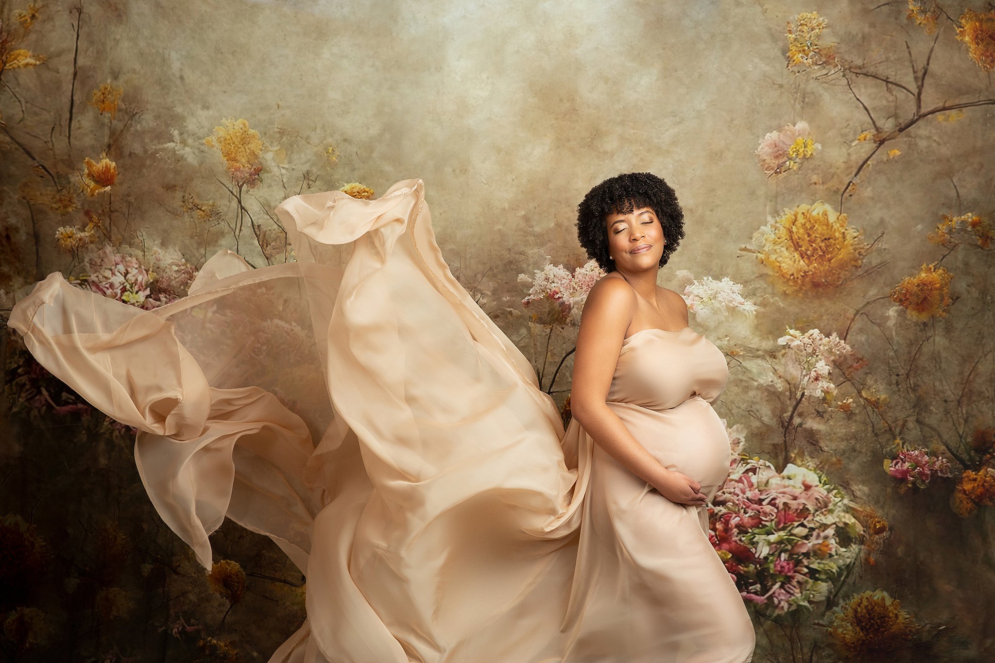 A mother to be in a beige maternity gown smiles over her shoulder with eyes closed