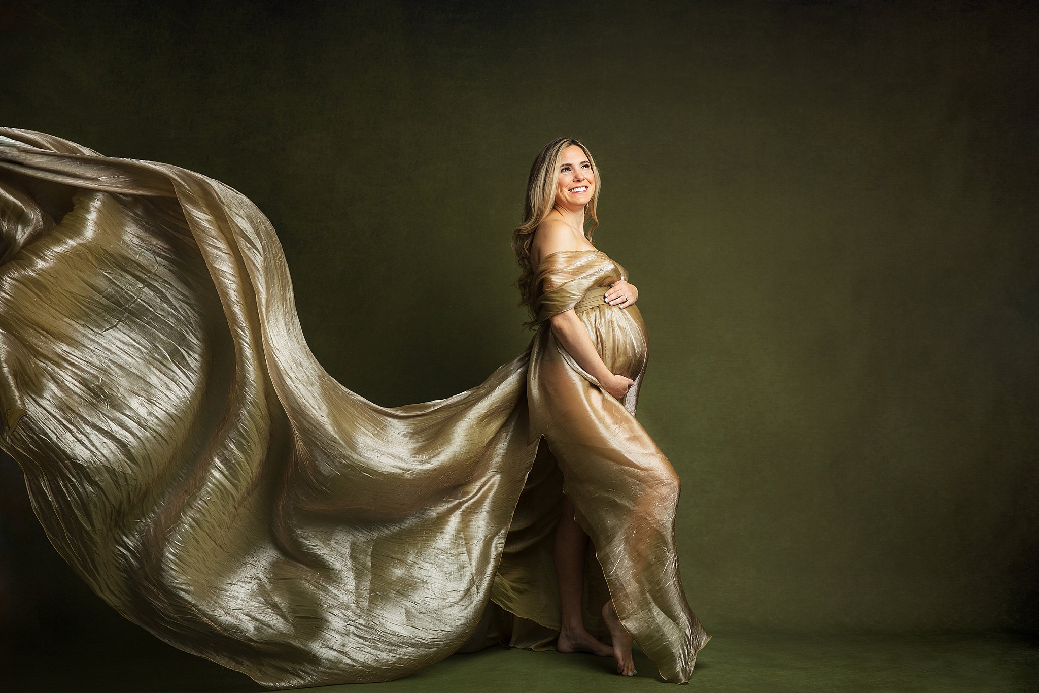 A mother to be stands in a studio in a gold maternity gown with a long flowing train behind her