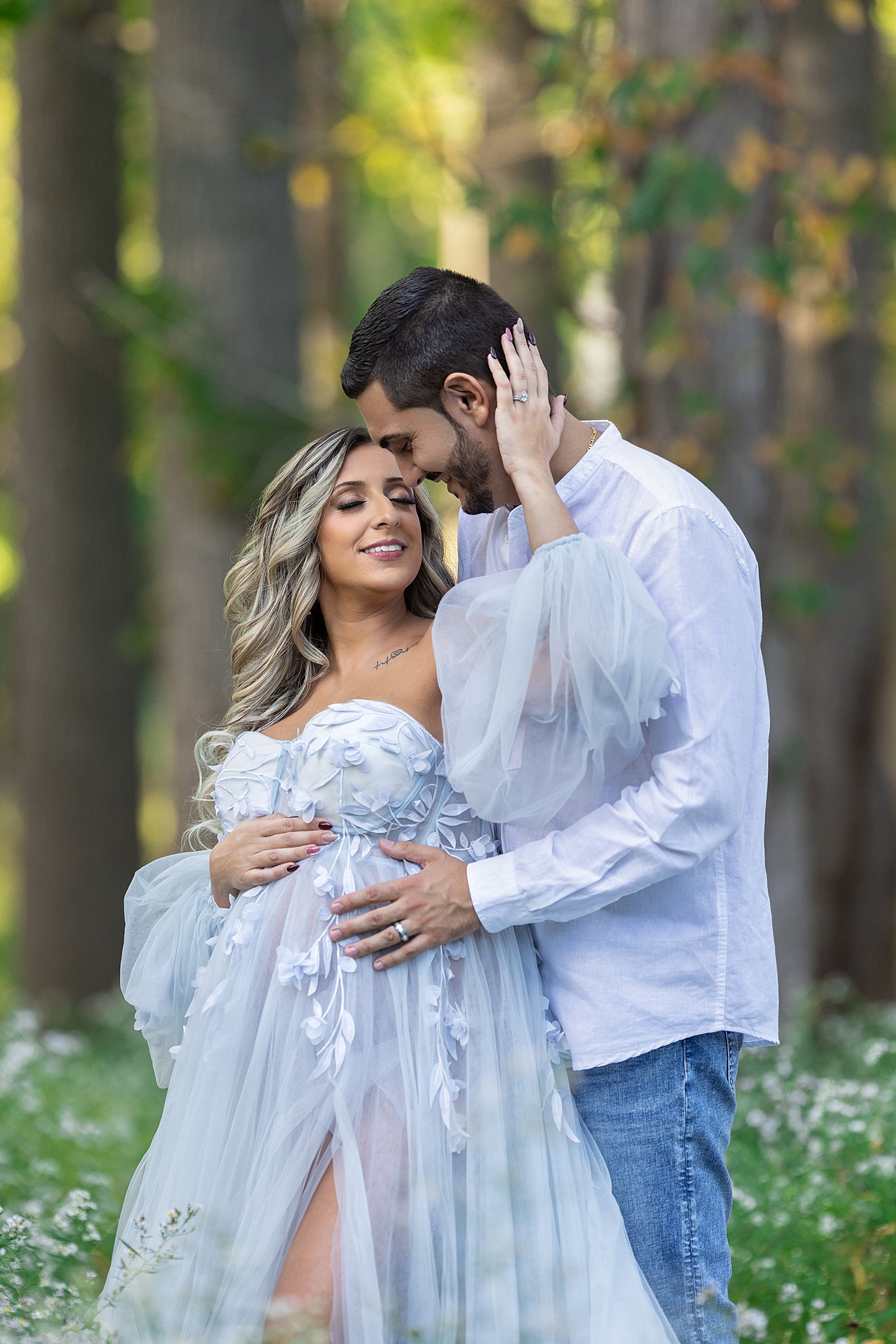 A mother to be in a blue maternity gown leans into and reaches up to her husband as they stand in a forest at sunset after visiting birthing centers nj