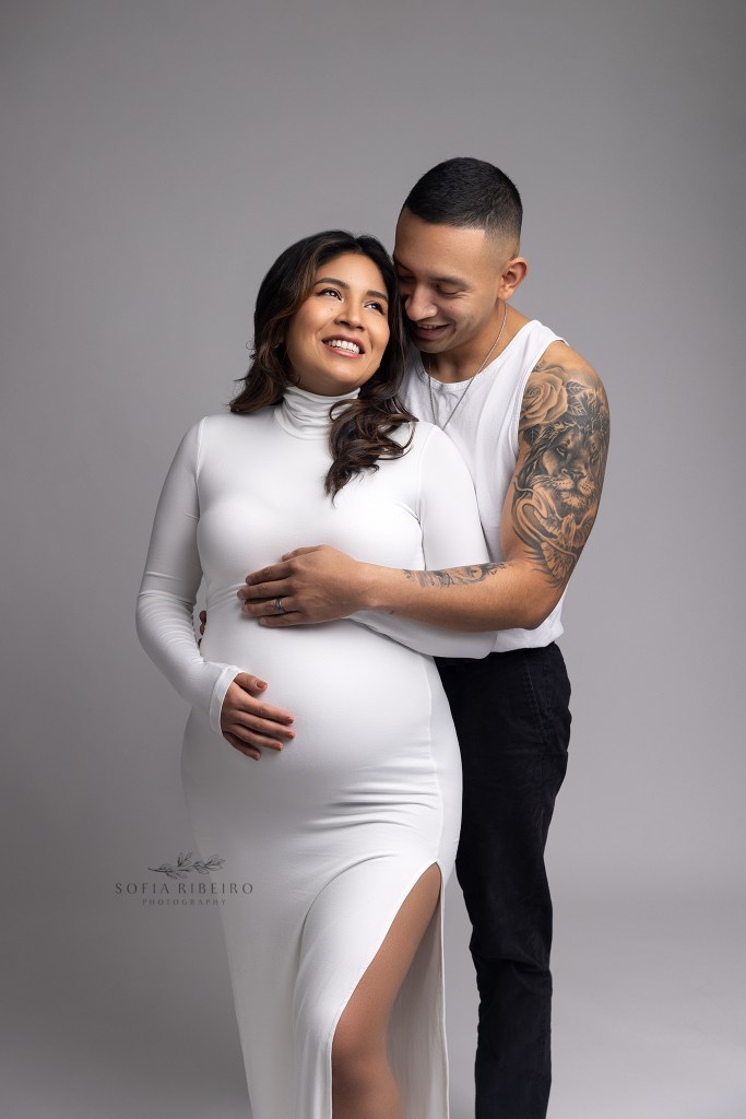 expectant mom leans against dad who is cradling her belly with his tattooed arm and smiling