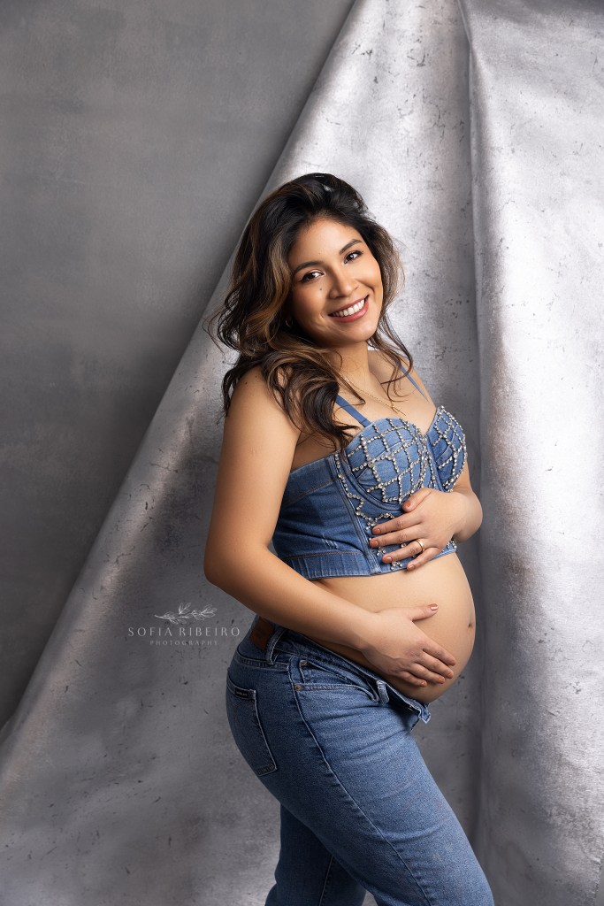 mom poses in a denim crop top and jeans against a silver backdrop during an nj maternity photo session