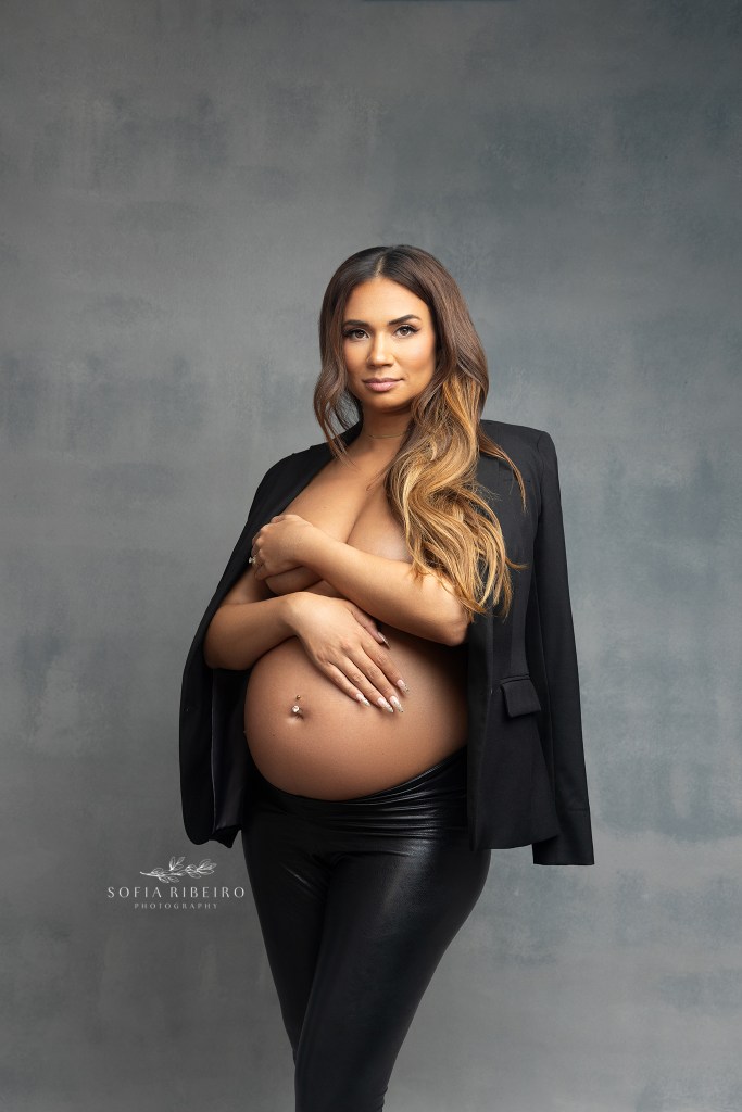 mom poses with attitude in an open black blazer on an apple box, with heels and leather tights during a maternity session in nj