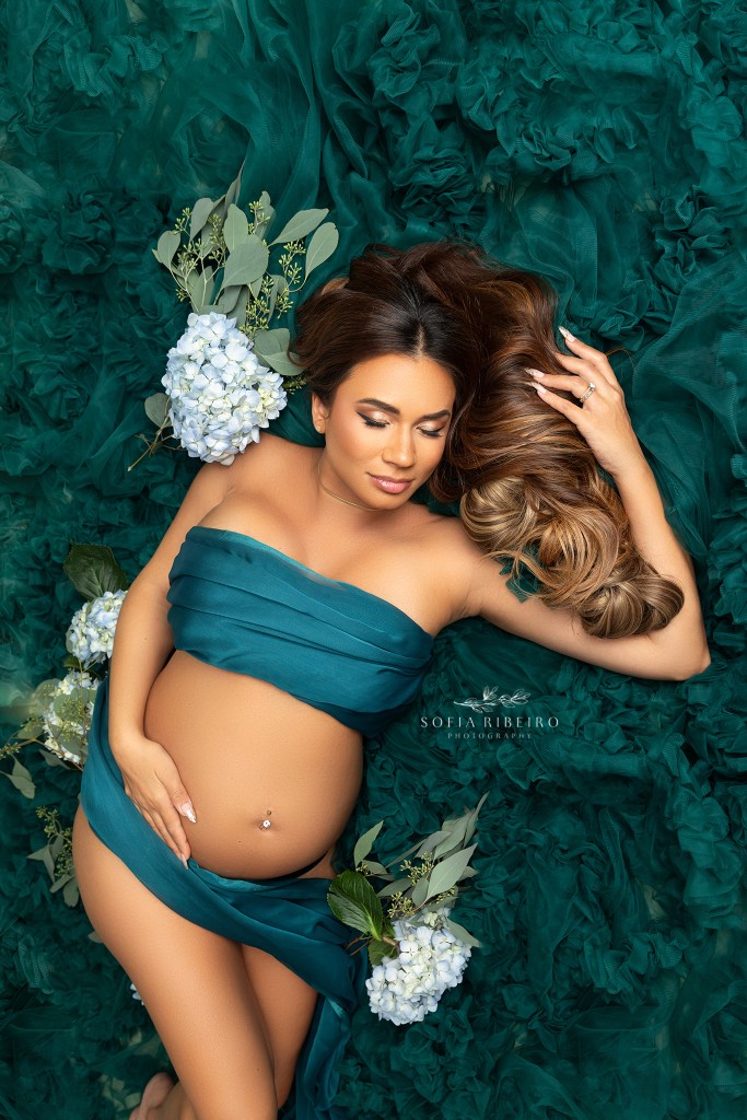 pregnant mom draped in green fabric nestled in tulle poses like a mermaid, exposing her belly during a maternity photo session in hoboken nj