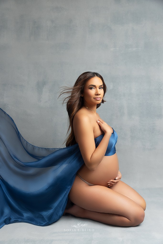 mom kneels on the floor with belly exposed, while blue fabric and hair blows back during a maternity session in hoboken nj