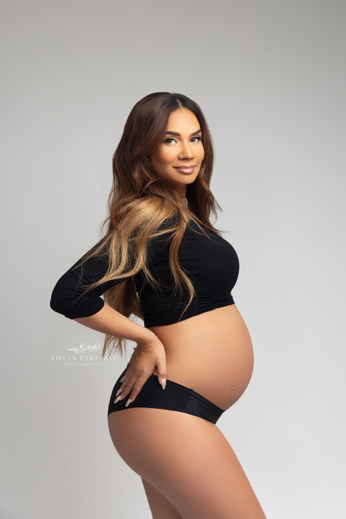 mom poses with hands on hips in a simple black drop top and black underwear while smiling at the camera during a maternity session in new jersey