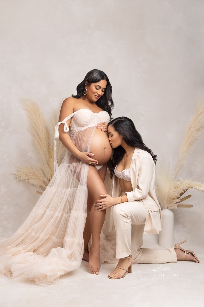 two moms pose together with one mom gently caressing mom's pregnant belly while wearing matching cream outfits.