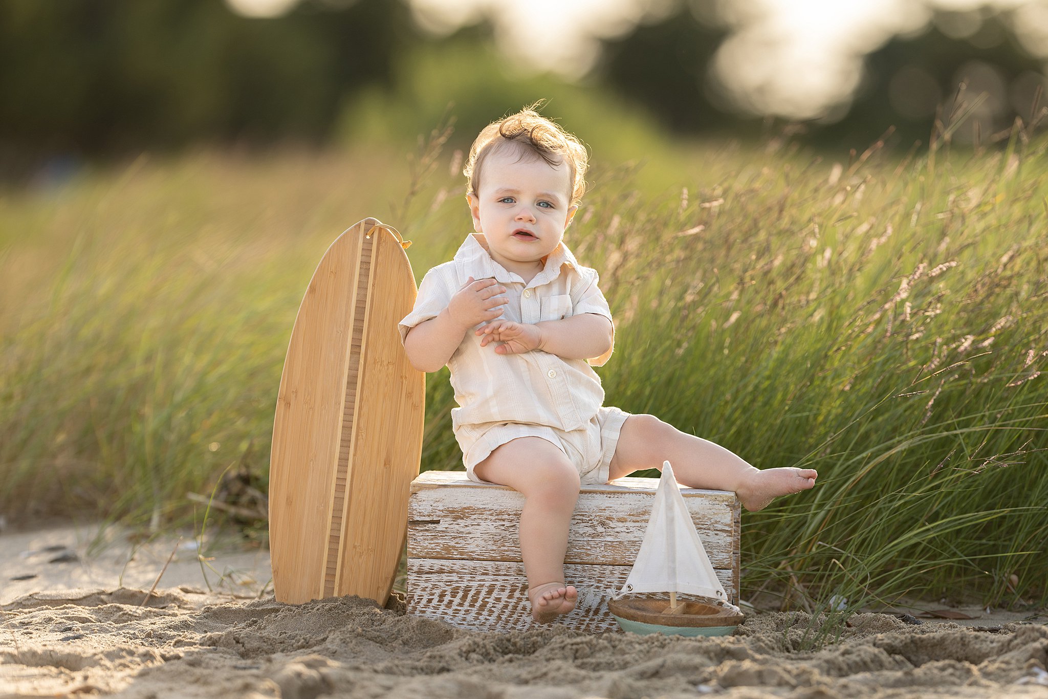 A young toddler boy sits on a wooden box on a beach at sunset next to a wooden sailboat and surfboard after visiting manymoons