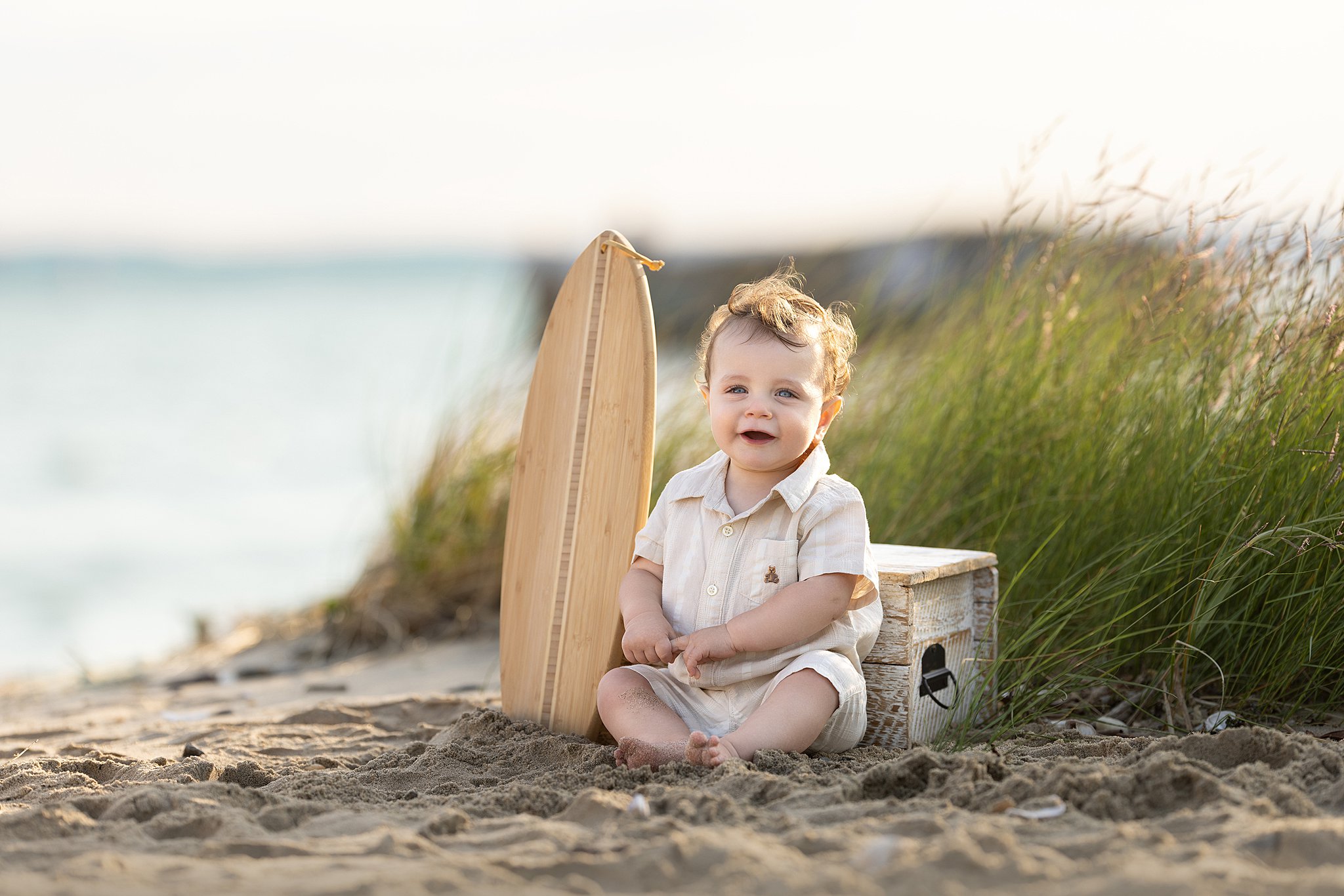 A toddler boy in a beige onesie sits on a beach next to a small wooden surfboard after meeting manymoons
