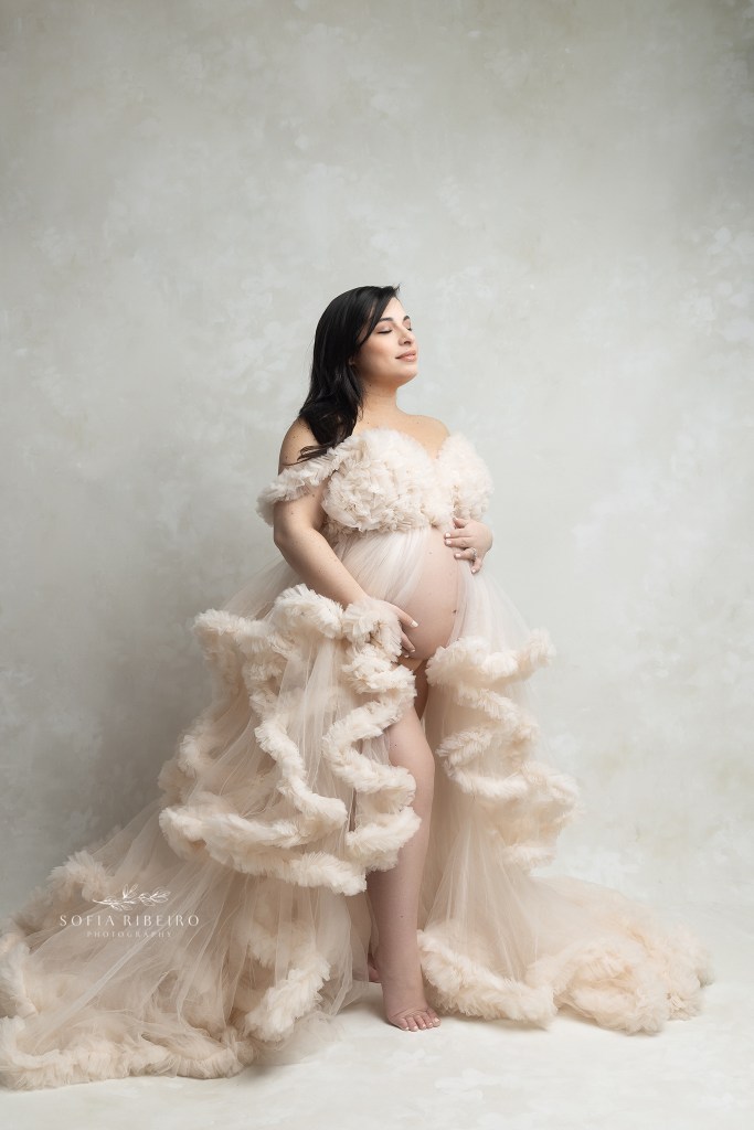 mom stands holding her belly in a cream ruffled gown exposing her belly during a maternity session in nj
