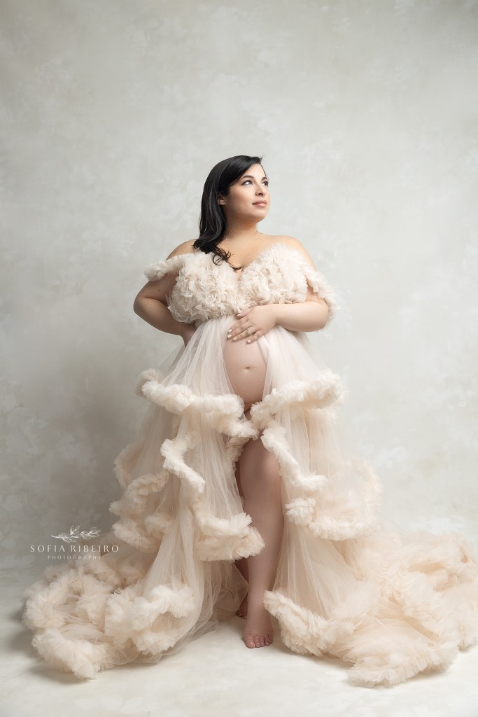 mom stands holding her belly in a cream ruffled gown exposing her belly during a maternity session in nj