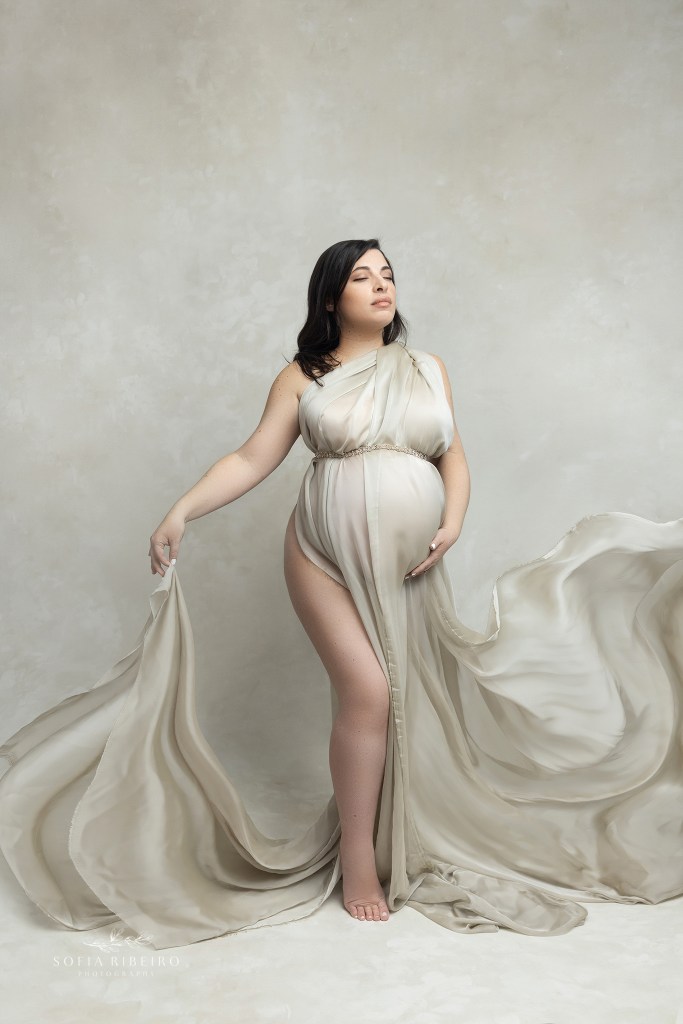 mom holds draped fabric in one hand and one under her belly while draped fabric blows softly around her pregnant belly.