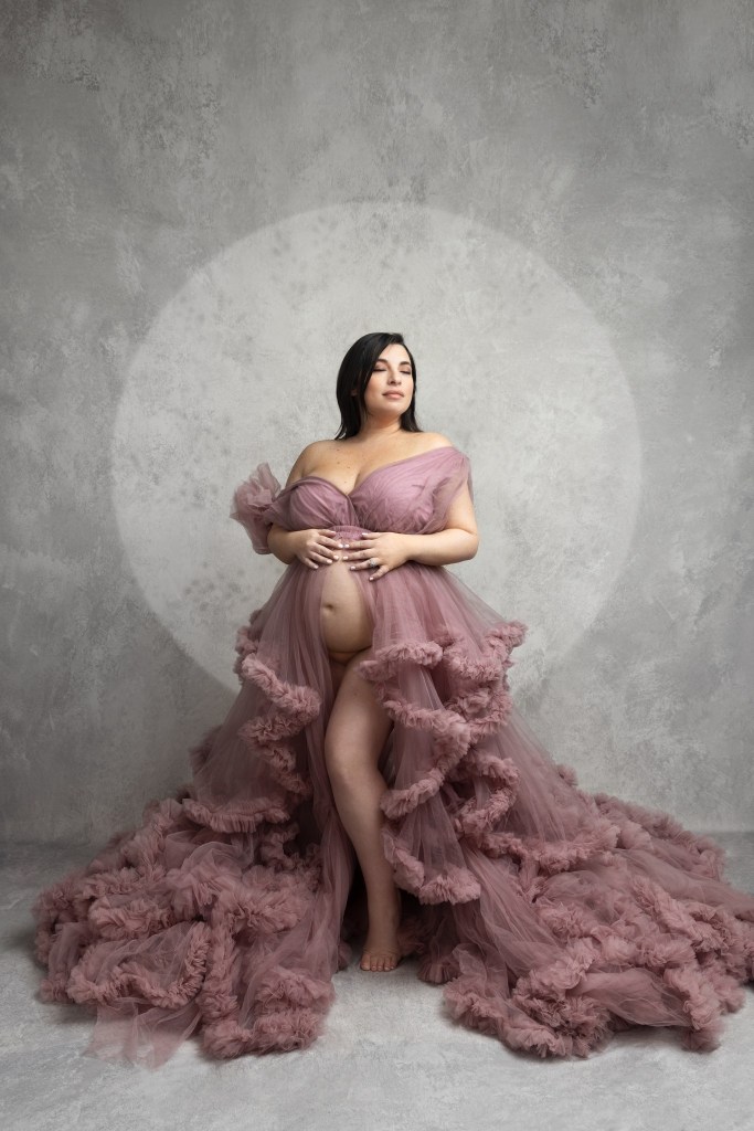 mom places both hands on her belly while looking over her shoulder in a pink maternity gown during a photoshoot in nj, surrounded by a halo of light