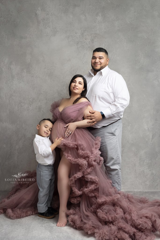 dad looks lovingly at expectant mom in a pink maternity gown for photos, while her son hugs her belly
