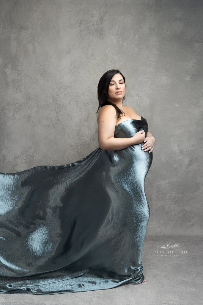 mom looks over her shoulder while draped in a belly hugging blue fabric being blown by a fan during a maternity session in nj