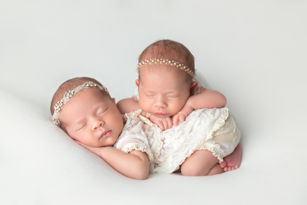 two baby twin girls are posed together by a nj newborn photographer in matching white outfits, while sleeping