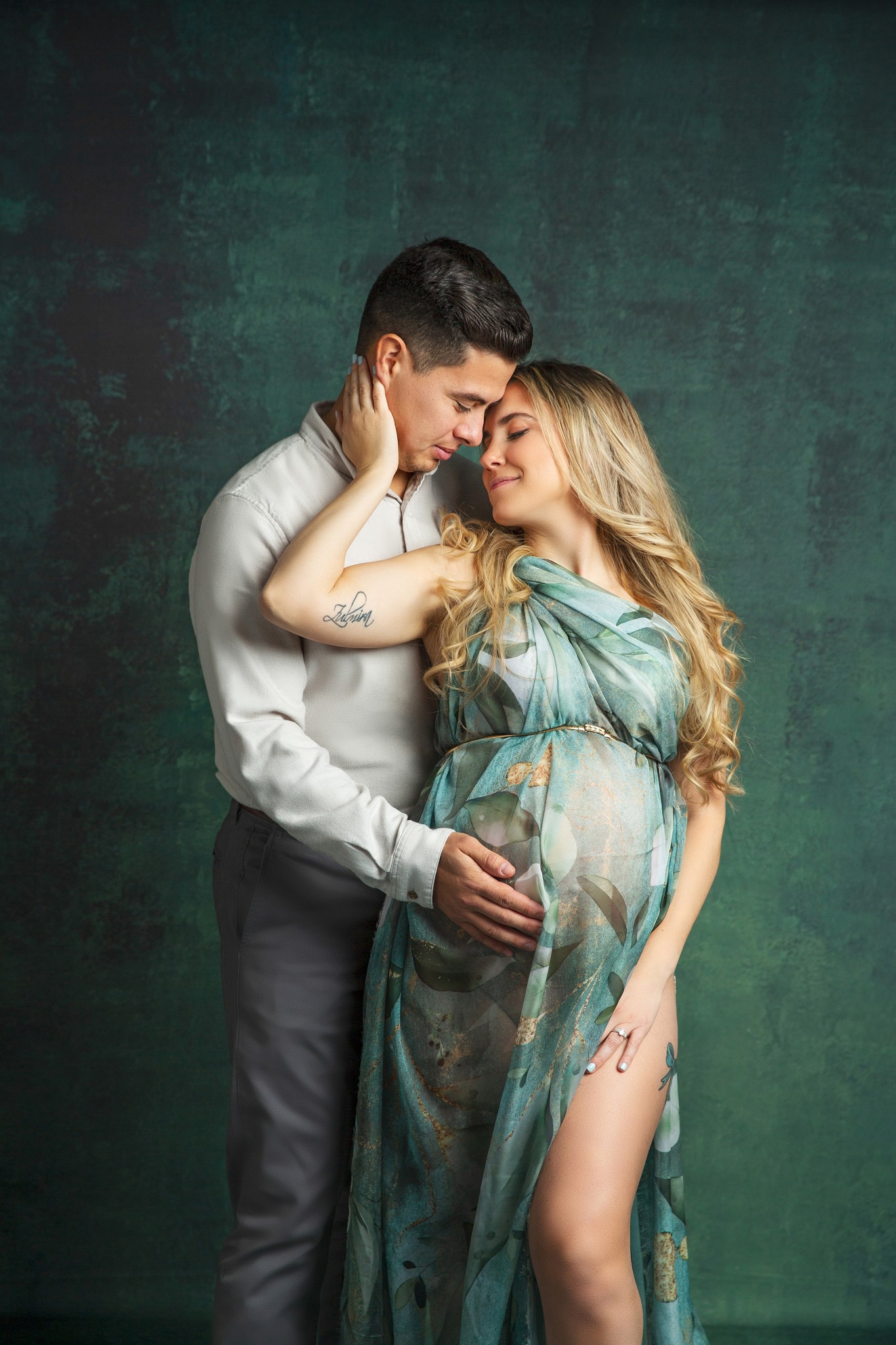 A mother to be in a green floral print dress leans into her husband who is holding the bump