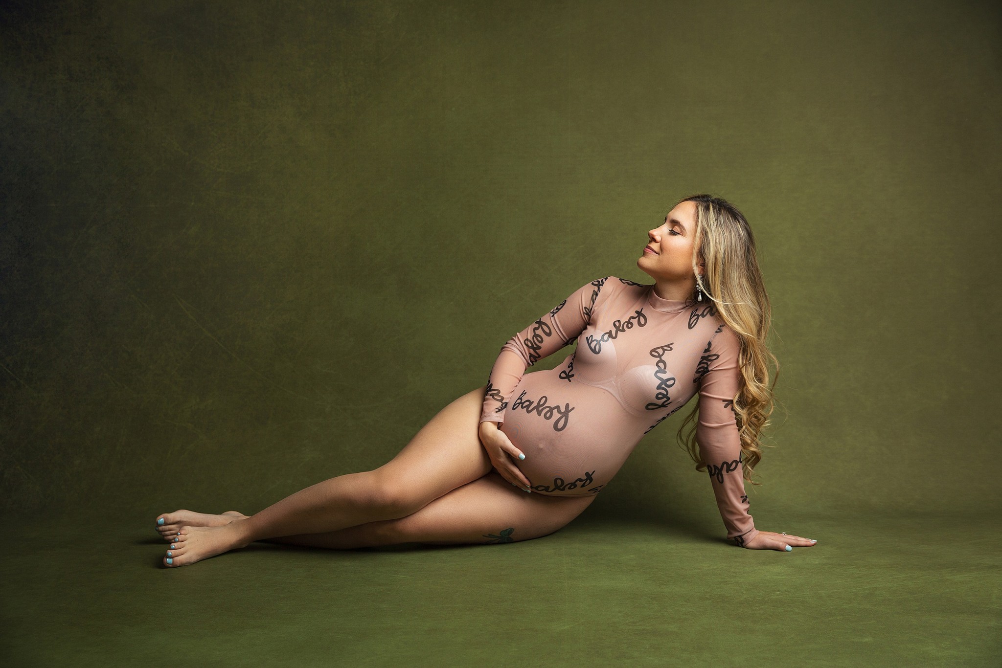 A mother to be lays across a studio floor with a hand on her bump in a sheer onesie after some prenatal yoga nj