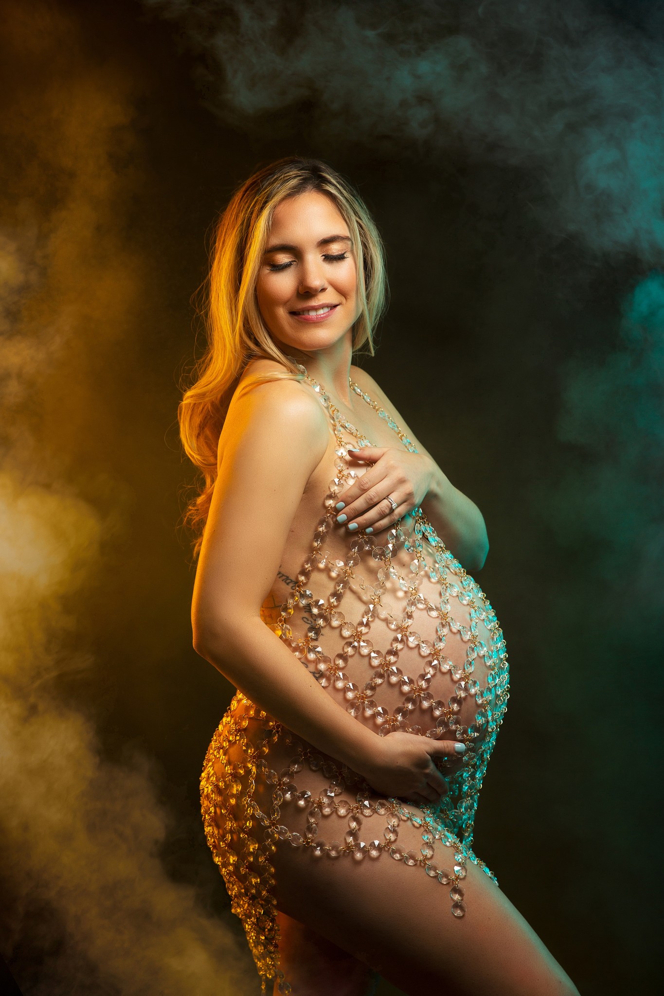 A mother to be in a glamorous maternity gown stands in a studio smiling down her shoulder after some prenatal yoga nj