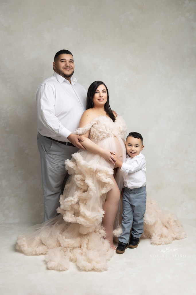 mom holds her little son's hands while her husband stands behind her dressed in a cream ruffled gown exposing her belly during a maternity session in nj