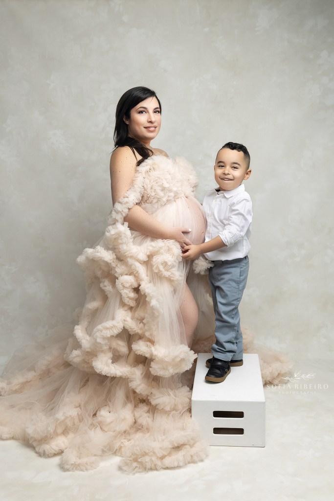 mom holds her little son's hands while dressed in a cream ruffled gown exposing her belly during a maternity session in nj